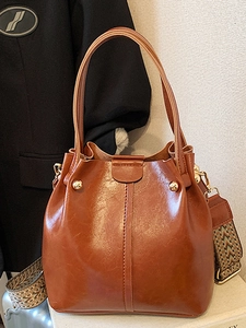 Glänzende braune Eimertasche mit weitem Ethno-Gurt und goldenen Nieten auf weißem Sockel