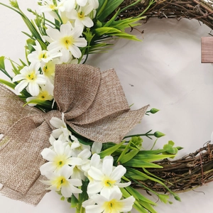 Easter cross wreath with white flowers, green leaves and a large burlap bow on a rattan base