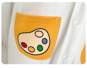 Close-up of white cotton baby romper featuring a yellow pocket with palette print and snap buttons