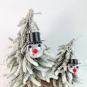 Close-up of disco ball snowman ornaments with black top hats on snowy Christmas trees