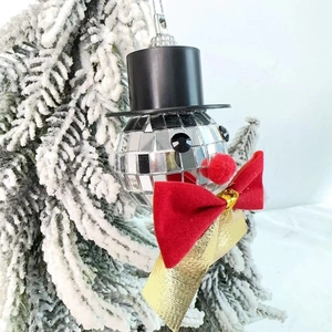 Close-up of a silver disco ball snowman ornament with red velvet bow on a snowy tree