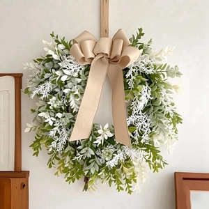 Detail of faux greenery wreath with white foliage and a large burlap bow on white wall