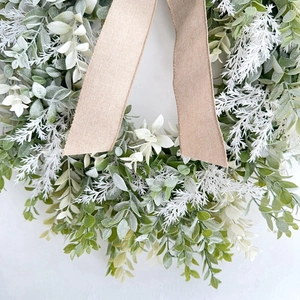 Artificial greenery wreath with white feathers and linen ribbon on a white wall