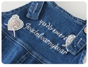 Details einer Baby-Latzhose aus Denim mit Herz-Spitzenstickerei und Perlen-Schleife