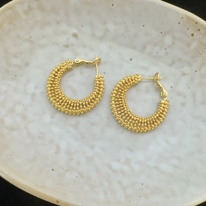 Close-up of golden C-shaped hoop earrings with woven bead texture on ceramic plate