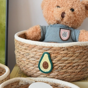Detail eines handgeflochtenen Strohkorbs mit Avocado-Häkelaufnäher und weißem Baumwollrand