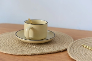 Close-up of a round woven jute placemat on a wooden table with a coffee cup and spoon