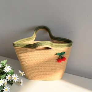 Close-up of woven straw tote bag featuring handmade red cherry crochet charms and green trim