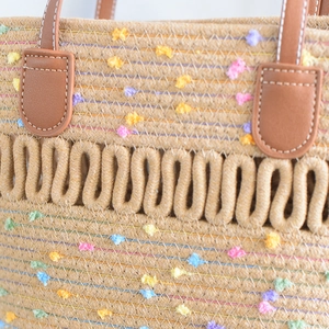 Close-up of woven tote bag with colorful pom-poms and brown leather handle stitching