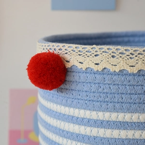 Close-up of woven cotton rope basket featuring blue stripes, lace trim and a red pompom