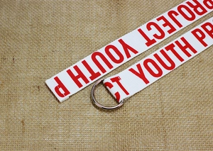 Détail d'une ceinture en toile blanche avec texte Project Youth rouge et boucle en D argent