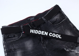 Détail de ceinture en toile avec imprimé HIDDEN COOL sur jean noir avec œillets métal
