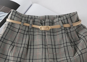 Detail of a thin beige leather belt with gold double-ring buckle on plaid grey trousers