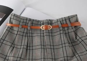 Close-up of a slim brown leather belt with gold oval buckle on grey plaid fabric