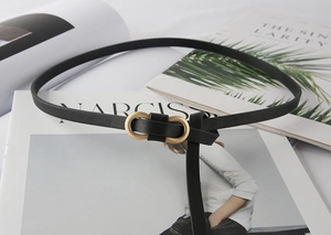 Detailed view of slim black leather belt with matte gold double ring buckle on a magazine