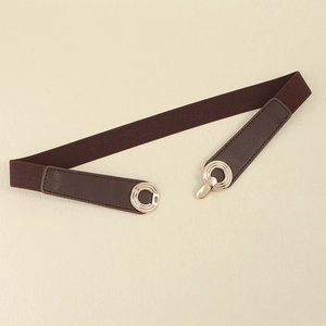 Detail of slim dark brown elastic belt featuring gold round metal buckle on beige surface