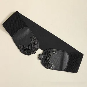 Détail de la ceinture élastique noire large avec boucle métallique florale sur fond beige clair