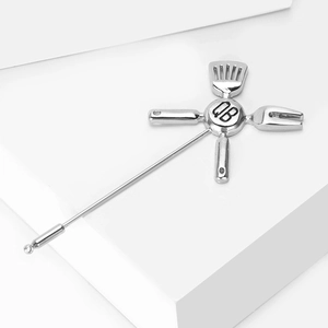 Macro shot of polished silver enamel brooch featuring kitchenware tools and QB engraving