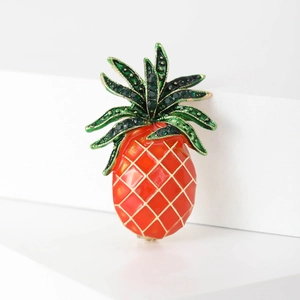 Close-up of a pineapple brooch with orange enamel and green crystals on white props