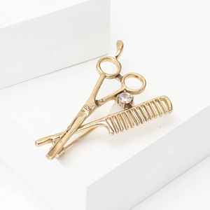 Broche coiffeur dorée avec ciseaux, peigne et zircon sur support blanc minimaliste