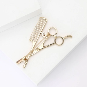 Détail de la broche dorée ciseaux et peigne avec cristal sur un piédestal blanc épuré