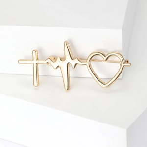 Détail de la broche EKG dorée avec croix et cœur ajouré sur un bloc minimaliste blanc pur