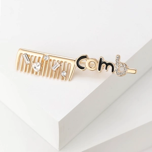 Golden comb brooch with white crystals and black enamel 'Comb' letter design