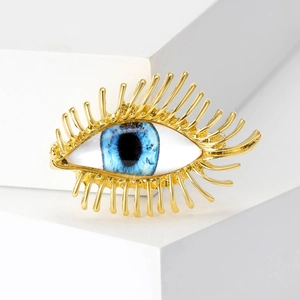 Détail de la broche œil avec cils dorés et iris bleu sur un présentoir blanc
