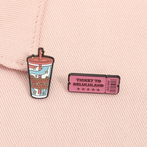 Detail of pink Delulu Juice and Delululand ticket enamel pins on fabric background