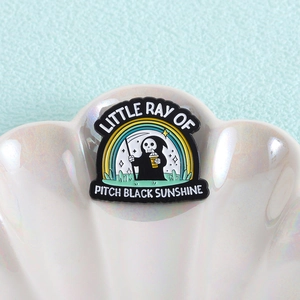 Détail du pin's faucheuse avec texte Little Ray of Pitch Black Sunshine sur plateau coquille
