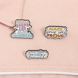 Détail de trois pin's émaillés aux couleurs pastel avec citations motivantes