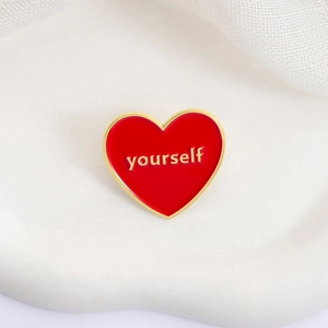Red heart enamel pin with gold 'yourself' text on a white ceramic plate