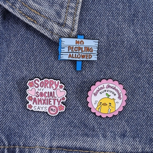 Three enamel pins featuring social anxiety themes pinned on a blue denim background fabric
