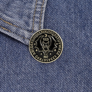Gothic bat enamel pin with Ouija board design and mystical symbols on blue denim
