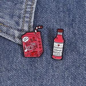 Close-up of enamel pins on denim, featuring Self Love juice box and sauce bottle designs