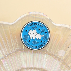 Detailed close-up of a blue enamel cat pin with crafting tools in a shell tray