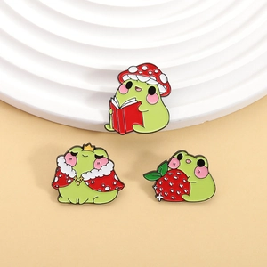 Macro detail of three enamel pins featuring frogs with mushroom, crown, and strawberry