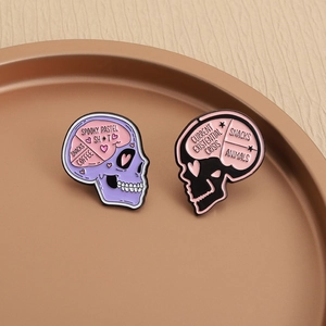 Detail of two pastel goth skull enamel pins with spooky humorous text in a metal tray