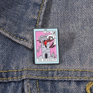 Detail of The Patriarchy tarot card enamel pin on denim fabric with XVI roman numeral