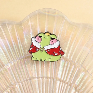 Close-up of cute frog prince enamel pin with red strawberry cloak in a shell tray