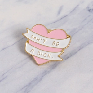 Pink heart enamel brooch with 'Don't Be A Dick' ribbon slogan on marble background