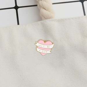Close-up of pink heart enamel pin with "Don't be a dick" text on a canvas bag