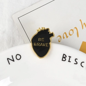 Broche cœur anatomique en émail Be Brave avec lettrage doré sur fond décoratif blanc