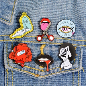 Set of 6 surrealist enamel pins including melting clock and heart eye on denim jacket
