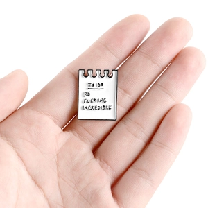 Handheld photo of an enamel notebook-shaped brooch with to-do list text