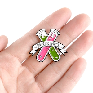 Emaille-Pin mit Reagenzgläsern, rosa und grüner Flüssigkeit, von Hand vor weißem Hintergrund gehalten