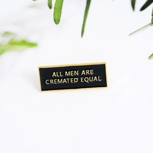 Black enamel brooch with 'ALL MEN ARE CREMATED EQUAL' text and golden alloy frame