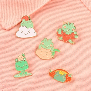 Set of five cute strawberry frog enamel pins displayed on a pink textured fabric background