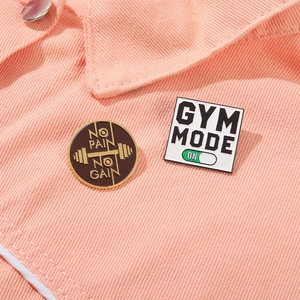Detail shot of No Pain No Gain and Gym Mode enamel pins on pink denim fabric surface