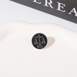 Detailed view of a minimalist black Libra scale enamel brooch on a white texture tray